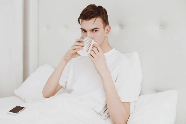 Handsome Young Man Sits Bed White Bedroom Drinks Coffee — Stock Photo, Image
