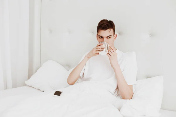 Handsome Young Man Sits Bed White Bedroom Drinks Coffee — Stock Photo, Image