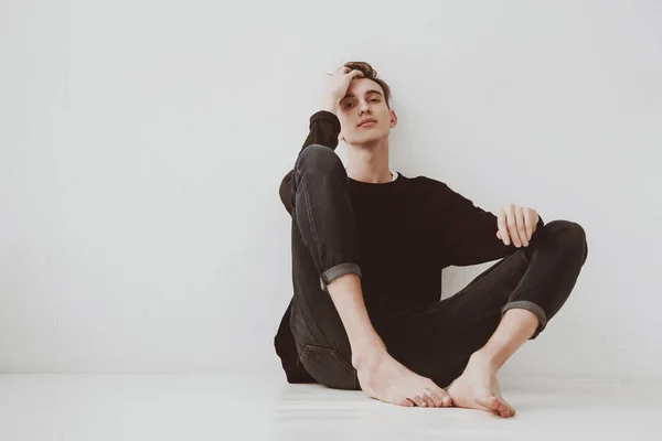 A young man, a student sits on the floor, against a white wall background, a pensive look. The guy is a model.
