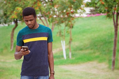 Siyah bir adam, parkta dikilen bir adam, sokakta, elinde cep telefonu, akıllı bir telefon. Telefondaki haritayı, iletişimi, yazışmaları incelemek için. Bilgi edinin. Millenial, öğrenci.