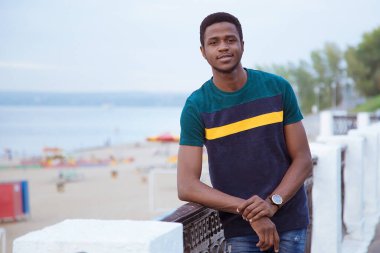 Siyahi bir adam, dirsek egzersizi yapıyor. Nazik, dostane bir gülümseme. Toprak seti, sahil, nehir denizi. Değişim öğrencisi. Uzayı kopyala Milenyum. Saat onun elinde..