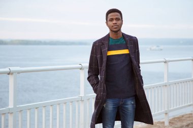 Siyah bir adam, ceketli bir adam, rıhtımda bir pedagog, nehrin arka planında, suda, denizde. Modaya uygun bir görünüş, imaj. Milenyum, Y Jenerasyonu