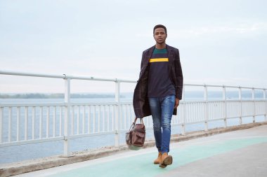 Paltolu siyah bir adam, rıhtımda bir pedagog, nehrin arka planında, su, deniz. Modaya uygun bir görünüş, imaj. Modern moda. Elinde bir çanta, elinde bir turist..