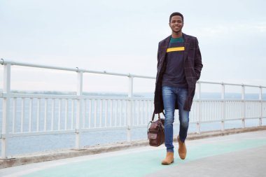 Paltolu siyah bir adam, rıhtımda bir pedagog, nehrin arka planında, su, deniz. Modaya uygun bir görünüş, imaj. Modern moda. Elinde bir çanta, elinde bir turist..