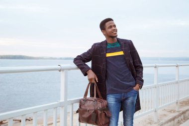 Paltolu siyah bir adam, rıhtımda bir pedzak, nehrin arka planında. Modaya uygun bir görünüş, imaj. Elinde bir çanta, elinde bir turist tutuyor. Korkuluklardan ameliyat et..