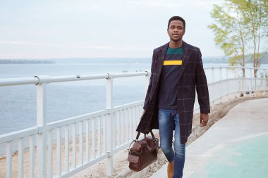 Paltolu siyah bir adam, rıhtımda bir pedagog, nehrin arka planında, su, deniz. Modaya uygun bir görünüş, imaj. Modern moda. Elinde bir çanta, elinde bir turist..