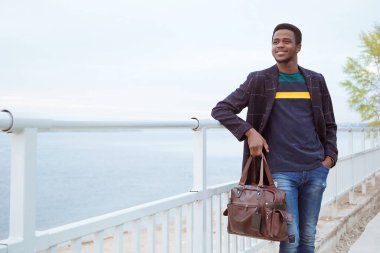 Paltolu siyah bir adam, rıhtımda bir pedzak, nehrin arka planında. Modaya uygun bir görünüş, imaj. Elinde bir çanta, elinde bir turist tutuyor. Korkuluklardan ameliyat et..