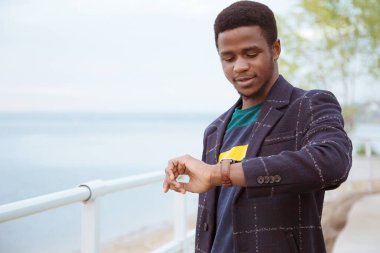 Paltolu siyah bir adam, rıhtımda bir pedzak, nehrin arka planında. Modaya uygun bir görünüş, imaj. Saatin akışını izliyor. Turist, değişim öğrencisi. Boşluğun kopyası.