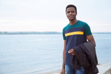 Siyah bir genç adam nehrin arka planında duruyor, elindeki deniz ceketini ve paltosunu tartıyor. Dört gözle bekliyorum. Geleceğe. Değişim öğrencisi, şık görünüşlü. Uzayın kopyası. Z Jenerasyonu.