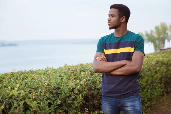 A black guy, a man stands against a river. Millenial, arms crossed. Against the background of the river, the sea. A thoughtful look, a copy of space. Closed gesture, keep for a distance. Careful.