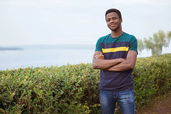 A black guy, a man stands against a river. Millenial, arms crossed. Against the background of the river, the sea. Copy space. Cheerful, smile.