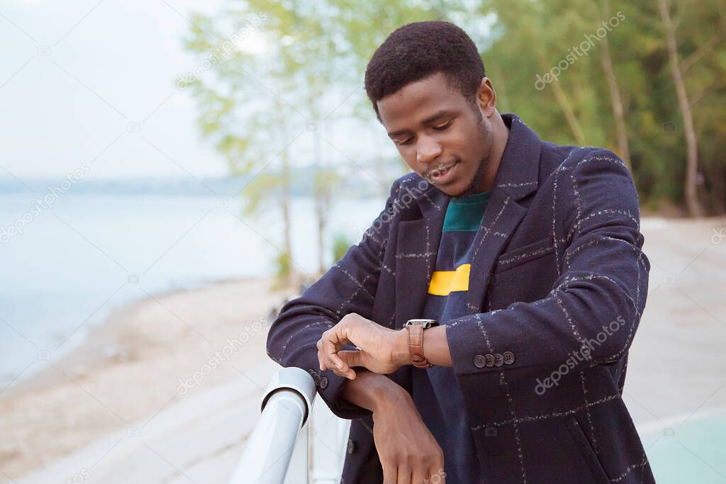 Black man in a coat, a pedzak on the waterfront, against the background of the river. Fashionable stylish look, image. Watches the clock on the hand. Tourist, exchange student. Copy of space.