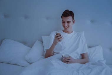 Genç yakışıklı bir adam yatakta uzanıyor ve telefona bakıyor, elinde bir akıllı telefon tutuyor. Adam gülüyor..