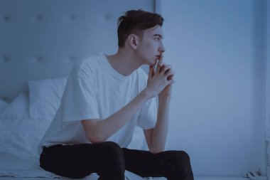 Beyaz tişörtlü, genç, yakışıklı bir adam yatağın köşesinde, yatak odasında oturuyor. Geceleri pencereden dışarı bakar.