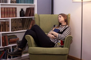 Güzel genç bir kadın elinde bir kitapla yeşil bir koltukta uzanıyor ve uyuyor, modern bir iç mekan. Kız çizgili bir bluz ve siyah bir korse giyiyor. Yaşam tarzı.