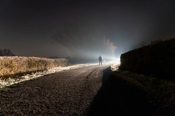 Gizemli, parlak bir ışığa karşı silueti olan bir adam, sisli bir kış gecesinde, kırsal bir yolda parlıyor. — Stok fotoğraf