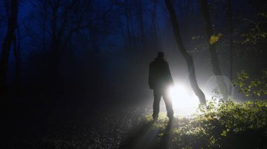 Kameraya arkası dönük bir adam ormanda parlayan bir ışığa bakıyor. Ürkütücü ve sisli bir kış akşamında.