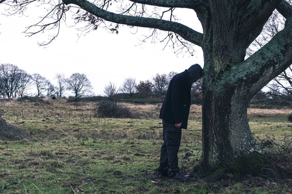Dziwny surrealistyczny obraz tajemniczego zakapturzonego mężczyzny uczącego się przeciwko drzewu w ponury, humorzasty dzień zimy na wsi — Zdjęcie stockowe