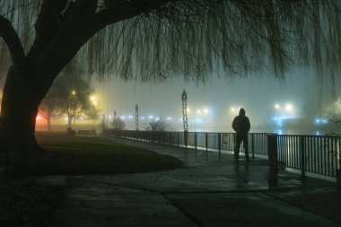 Sisli bir atmosferik kış gecesinde bir nehrin kenarında sokak ışıklarına bakan kapüşonlu gizemli bir siluet.