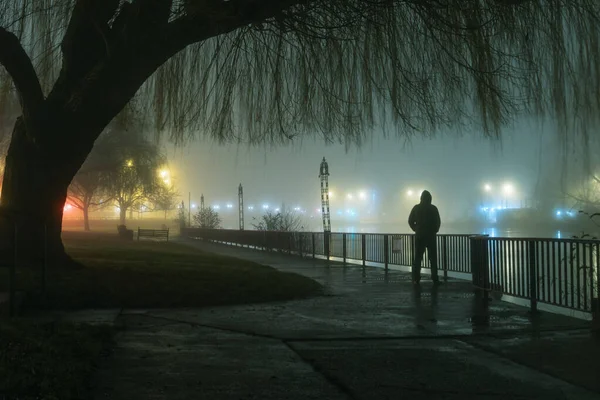 Une mystérieuse silhouette à capuchon humoristique silhouette contre les lampadaires par une rivière sur une nuit d'hivers atmosphériques brumeux — Photo