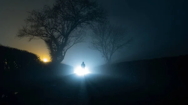 Korkunç, gizemli bir kukuletalı figür, kırsal bir şeritte parlak bir ışığın önünde duruyor, sisli bir kış gecesinde.. — Stok fotoğraf