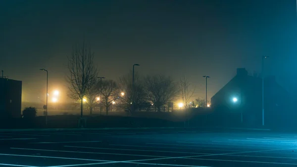 An empty car park with street lights glowing in the distance on a mysterious moody,  foggy atmospheric winters night — Stock Photo, Image