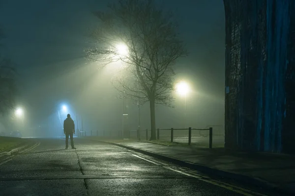 Een mysterieuze figuur bij een straatlantaarn van de stad op een humeurige, mistige sfeervolle winters nacht — Stockfoto