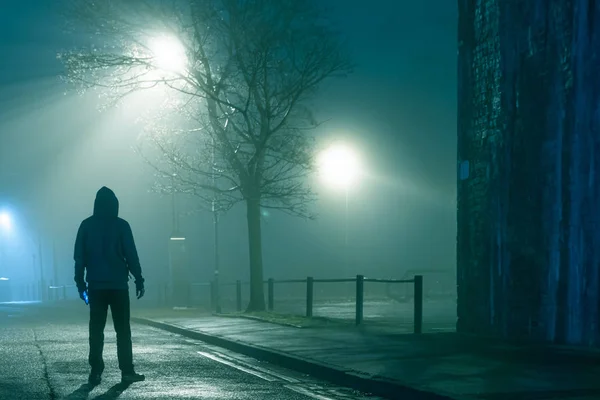 Таинственная фигура в капюшоне с телефоном, стоящая на городской улице. В капризный туманный зимний вечер — стоковое фото
