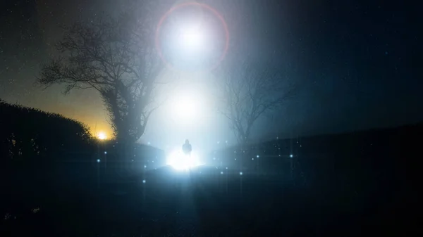 Um sobrenatural, conceito UFO de uma figura assustadora, misteriosa encapuzada, em pé na frente de uma luz brilhante no céu, em uma pista de campo, em uma noite assustadora de invernos nebulosos . — Fotografia de Stock