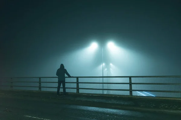 A hooded figure, standing with back to camera on a bridge, looking out at street lights. On a foggy night.