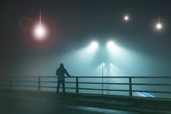 A hooded figure, standing with back to camera on a bridge, looking at UFO alien spaceships coming down from the sky Street lights. On a foggy night.
