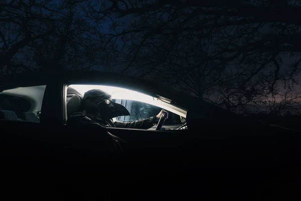 A sinister hooded figure wearing a plague doctor mask inside a car. On a spooky country road on a winters night — Stock Photo, Image