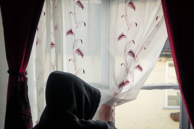Kapüşonlu bir figür, pencereden dışarı bakıyor. Akıl sağlığı ve izolasyon sorunlarını belirten.