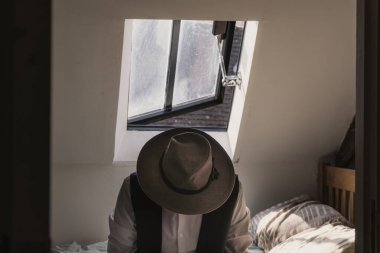 Fötr şapkası ve yeleği olan bir adam arka penceresi olan bir yatakta oturuyor. Klasik bir düzenleme ile