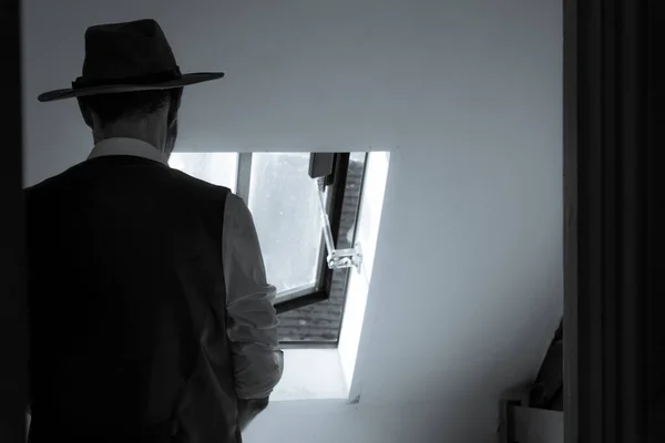 A man looking out of a window, back to camera, wearing a waistcoat and fedora hat. With a classic black and white edit