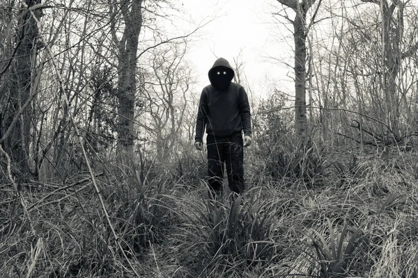 A scary, hooded figure with glowing evil eyes and blank black space where his face should be, standing in a forest in winter. With a vintage edit.