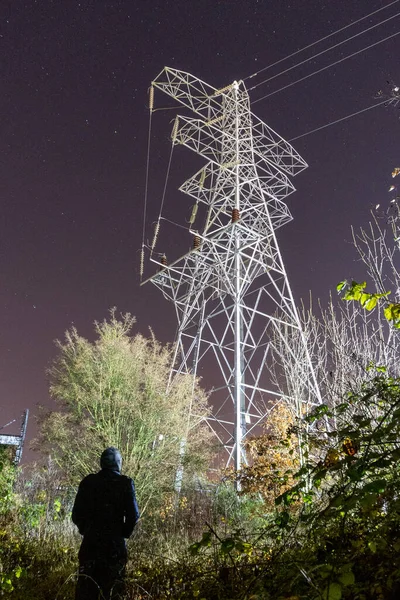 A hooded figure, back to camera. Looking up at a electricity pylon on a winters night.