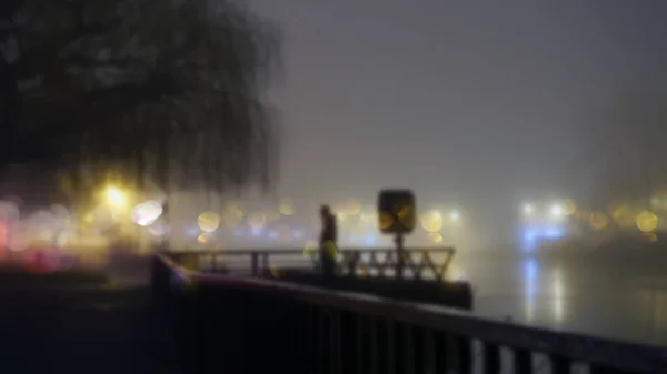霧深い冬の夜に川の隣の街灯のそばに立つ神秘的な姿 ぼんやりしたボケでフォーカス編集なしで — ストック写真