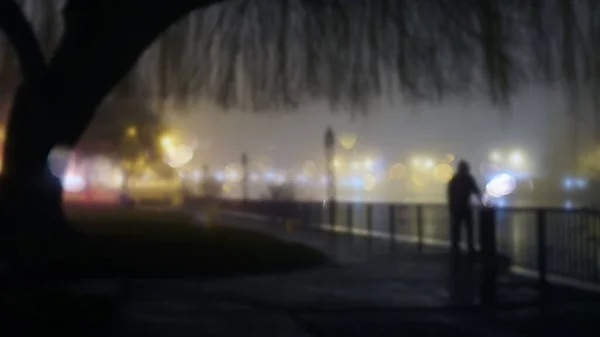 霧深い冬の夜に川の隣の街灯のそばに立つ謎のフード姿 ぼんやりしたボケでフォーカス編集なしで — ストック写真