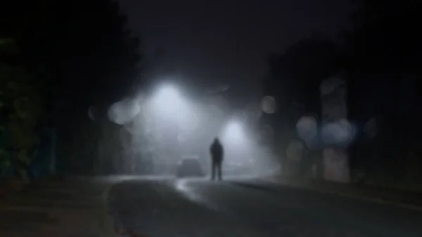 A mysterious figure standing in the middle of a road with street lights on a misty winters night. With a blurred, bokeh, out of focus edit