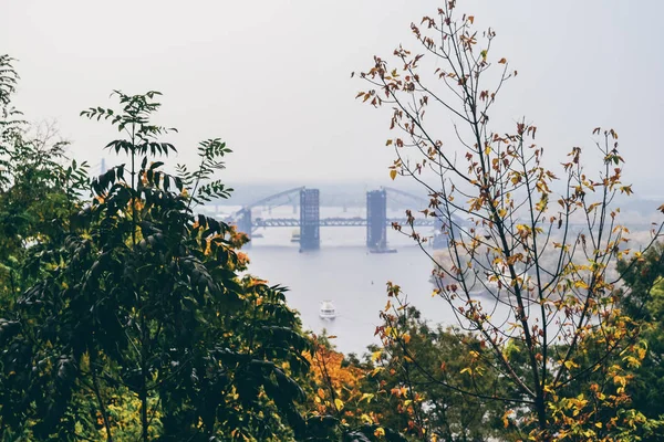 Kiev, ukraine - ca. Oktober 2011: herbstlicher Park mit Bäumen mit trockenen gelben und orangefarbenen Blättern mit Podilsko-voskresensky Brücke in der Ferne in kiev, ukraine cicra Oktober 2011. — Stockfoto