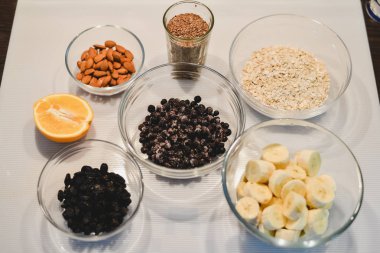 Bir sağlıklı şekersiz için malzemeler ve un ücretsiz muz yulaf ezmesi muffins: kesilmiş muz, böğürtlen, Kuru üzüm, badem, yulaf, bir cam kase ve bir yarım veya bir portakal beyaz bir yüzeye keten tohumu.