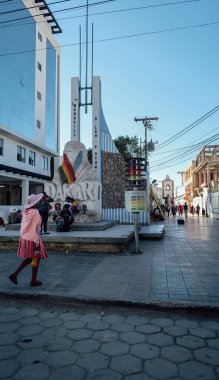 UYUNI, BOLIVIA - CIRCA NOVEMBER 2019: Geleneksel Bolivya elbiseli kadın Uyuni Meydanı 'ndaki Dakar mitingi için anıtın yanında yürüyor.