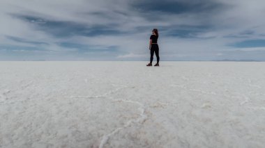 UYUNI, BOLIVIA - CIRCA NOVEMBER 2019: Siyah elbiseli kız bulutlu mavi gökyüzü ile güneşli bir günde Salar de Uyuni 'nin sonsuz boş tuz çölünün ortasında duruyor.