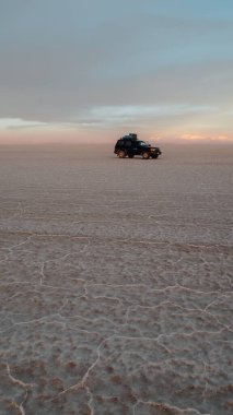 UYUNI, BOLIVIA - CIRCA NOVEMBER 2019: Salar de Uyuni 'nin sonsuz boş tuz çölünün ortasında bulutlu gökyüzü ile parlak mor günbatımında siyah SUV.