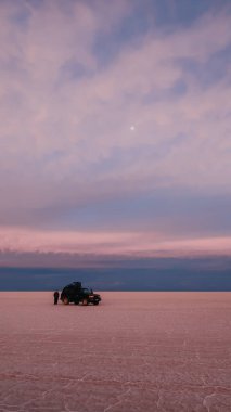 UYUNI, BOLIVIA - CIRCA NOVEMBER 2019: Salar de Uyuni 'nin sonsuz boş tuz çölünün ortasında bulutlu gökyüzü ile parlak mor günbatımında siyah SUV.
