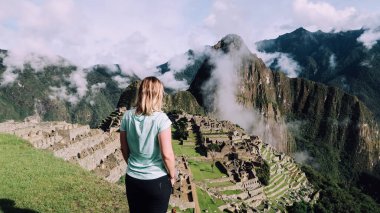 MACHU PICCHU, PERU - CIRCA Kasım 2019: Sarı saçlı kız dağları ve bulutları gören Machu Picchu 'nun ikonik manzarasının tadını çıkarıyor.