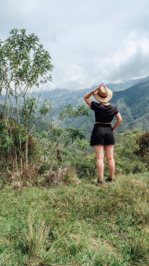 SANTANDER, COLOMBIA - CIRCA Aralık 2019: Şortlu ve kovboy şapkalı kız orman, yeşil tepeler ve bulutlu gökyüzü ile üzerinde dağların olduğu doğal manzaraya bakıyor.