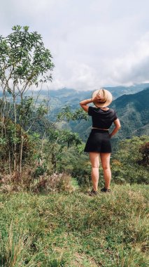 SANTANDER, COLOMBIA - CIRCA Aralık 2019: Şortlu ve kovboy şapkalı kız orman, yeşil tepeler ve bulutlu gökyüzü ile üzerinde dağların olduğu doğal manzaraya bakıyor.
