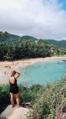TAYRONA, COLOMBIA - CIRCA Aralık 2019: Siyah elbiseli bir kız, güneşli bir günde Karayip denizi ve tropikal ormanların turkuaz suyuyla Cabo San Juan 'ın popüler plajının keyfini çıkarıyor.